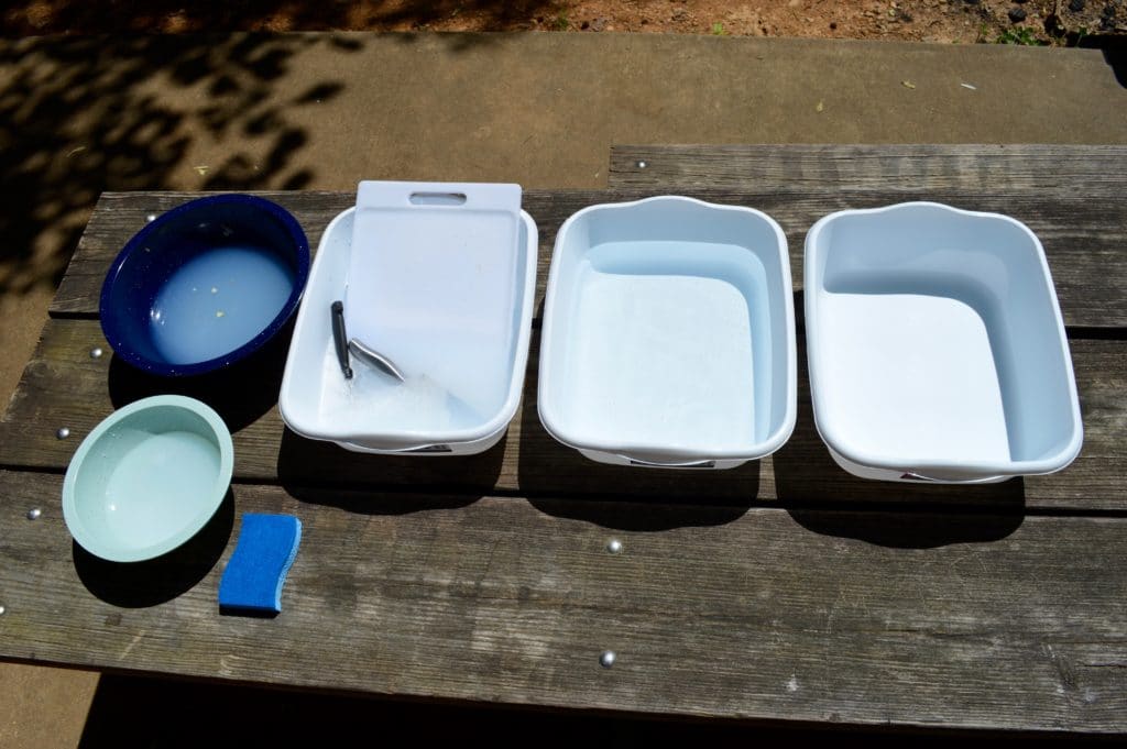 three dishpans with water, soap, and sanitizer set up on a picnic table ready to wash dishes while camping
