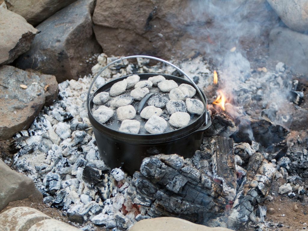 a dutch oven in a bed of hot coals with charcoal on the lid