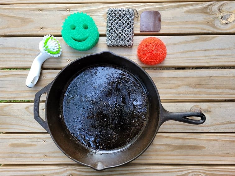 dirty cast iron skillet on a picnic table surrounded by cleaning tools 