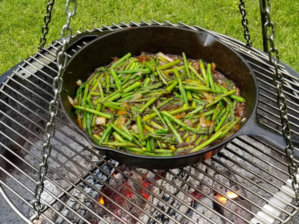 asparagus and chopped onions cooking in a cast iron skillet over a fire pit 