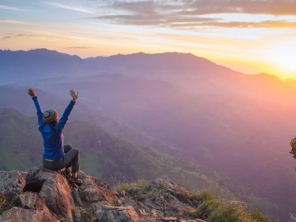 a woman sitting on a mountaintop with outstretched arms facing the rising sun.