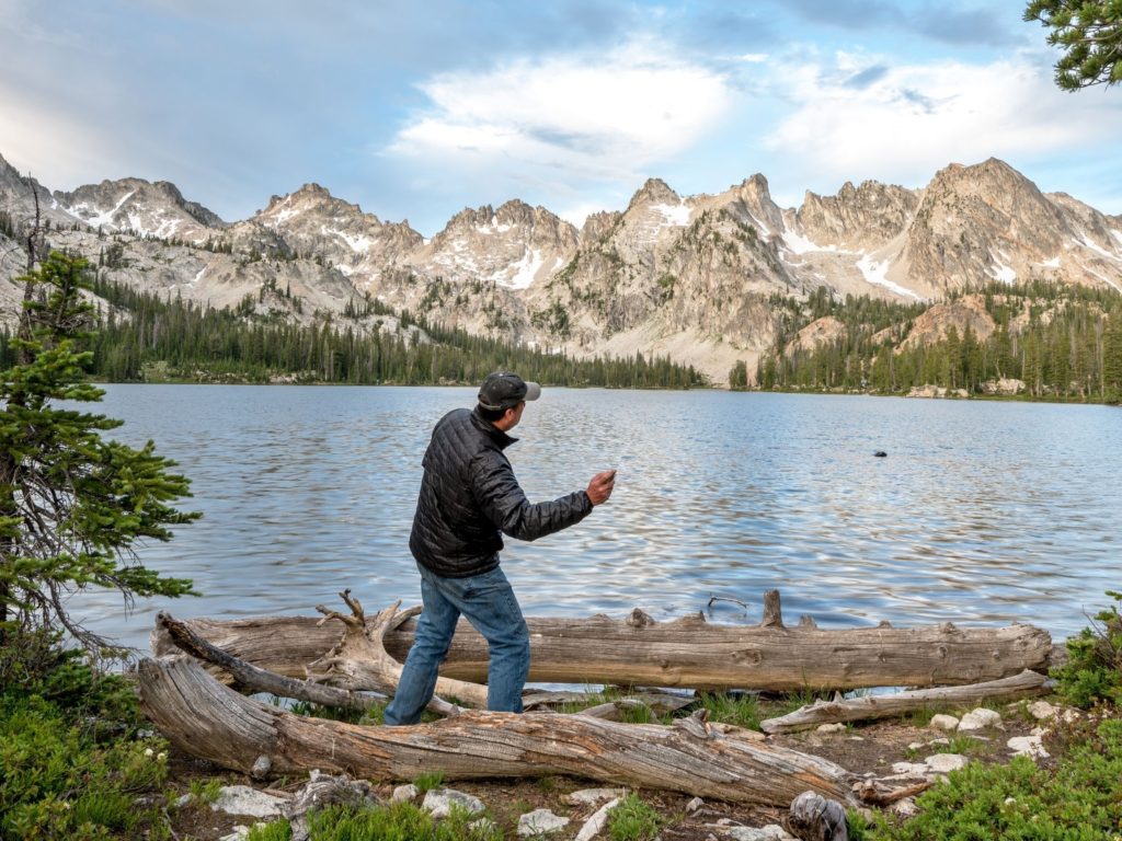 a man skipping rocks on a lake with beautiful tree-lined and snow-capped mountains in the background