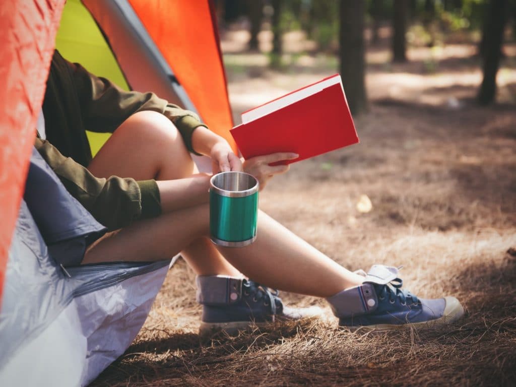 a woman sitting partially inside a camping tent reading a book and holding a coffee mug