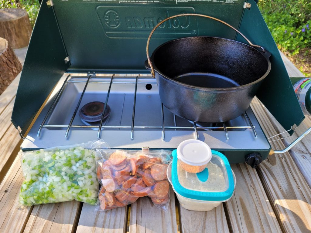 cast iron dutch oven on a camp stove with bagged recipe ingredients on a picnic table. 