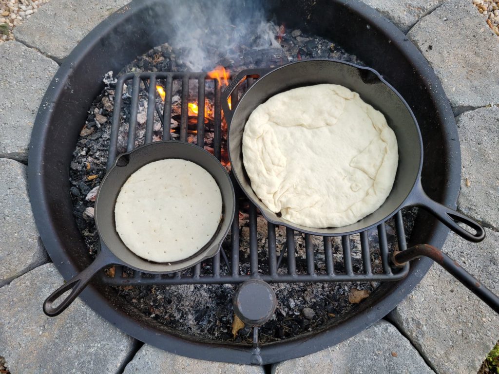 two pizza crusts cooking in cast iron skillets over a campfire